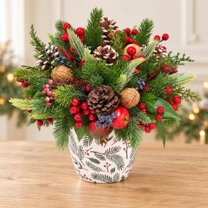 Könnte beinhalten: Ein Blumenarrangement in einem weißen Keramiktopf mit grünen Tannenzweigmustern. Der Strauß besteht aus grünen Tannennadeln, braunen Tannenzapfen, Walnüssen, roten Beeren und kleinen roten Äpfeln in einer dichten Anordnung.