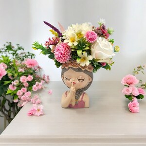 May include: A ceramic planter shaped like a girl with pink and white flowers in her hair. The planter is holding her finger to her lips as if to say "shhh".
