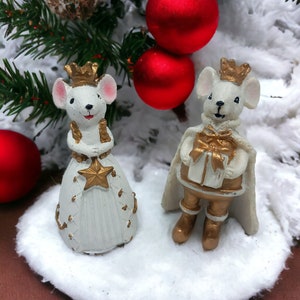 Puede incluir: Figuras de ratones blancos vestidos de rey y reina, de pie sobre una base nevada con adornos navideños rojos y una rama de pino verde en el fondo.