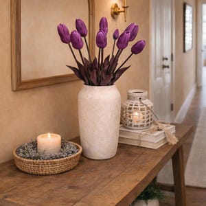 May include: A wooden console table features a white vase filled with purple tulips, a lit candle in a woven basket, and a decorative lantern. A mirror and wall sconce are in the background, creating a warm atmosphere.