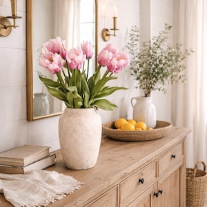 May include: A wooden cabinet with drawers and doors holds a vase of pink tulips. A gold-framed mirror hangs above the cabinet, reflecting the flowers. A woven tray with lemons and a white vase with greenery are also on the cabinet.