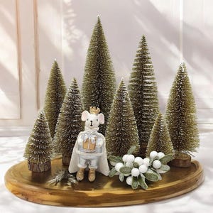 Könnte beinhalten: Eine weiße Mausfigur mit Krone und einem Geschenk in der Hand sitzt vor einer Gruppe von sechs kleinen, flockigen Weihnachtsbäumen auf einem Holztablett. Die Bäume sind alle unterschiedlich groß und mit weißen Beeren verziert.