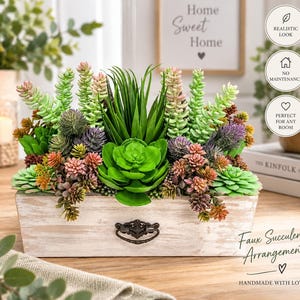 以下が含まれることがあります： 緑、ピンク、紫の様々な植物をあしらった人工多肉植物のアレンジメント。アンティーク調の白い木製長方形プランターに入っており、前面中央にはダークメタルの引き出し用取っ手が付いています。