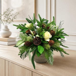 Op de afbeelding: Een bloemstuk in een grijze, getextureerde pot met crèmekleurige rozen, donkerbruine dennenappels, gedroogde lotuspeulen en diverse groene, dennenachtige bladeren. Het arrangement bevat kleine witte bessen en lange, smalle groene bladeren.