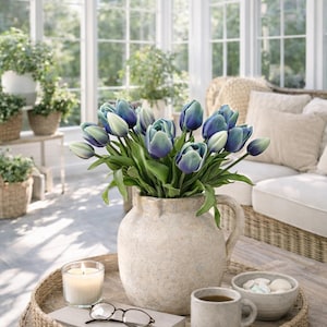 May include: A tranquil interior scene with a woven tray holding a vase of blue and white tulips, a lit candle, reading glasses, a book, a coffee mug, and a small bowl. The room has large windows and a neutral colour palette.