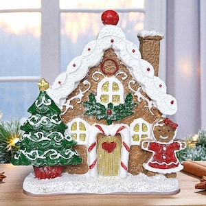May include: A festive gingerbread house decoration with a green Christmas tree and a gingerbread person. The house features white icing, red accents, and a red ball on top. The base is covered in white glitter, creating a wintery scene.