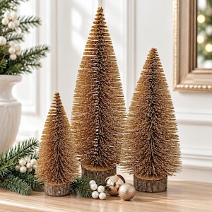 May include: Three decorative bottle brush trees in varying heights with metallic gold bristles and natural wood slice bases. The trees are arranged with small white berry sprigs and two gold spherical ornaments on a flat surface.