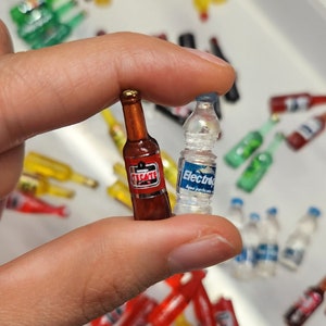 May include: A close-up of a hand holding two miniature bottles. The bottle on the left is brown glass with a red label that says "Tecate". The bottle on the right is clear glass with a blue label that says "Electropura Agua Purificada". The bottles are surrounded by a variety of other miniature bottles in different colors and shapes.