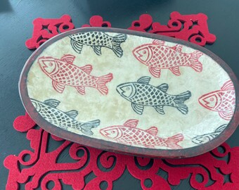 Puede incluir: Un plato de servir ovalado con fondo blanco y un patrón de peces rojos y negros. El plato tiene un borde marrón y está sobre una alfombra de fieltro roja.