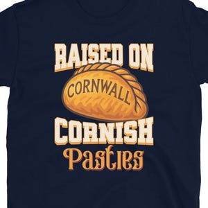 May include: Dark blue t-shirt featuring the text "RAISED ON CORNWALL CORNISH Pasties" in white and orange. The design includes a graphic of a golden-brown Cornish pasty.
