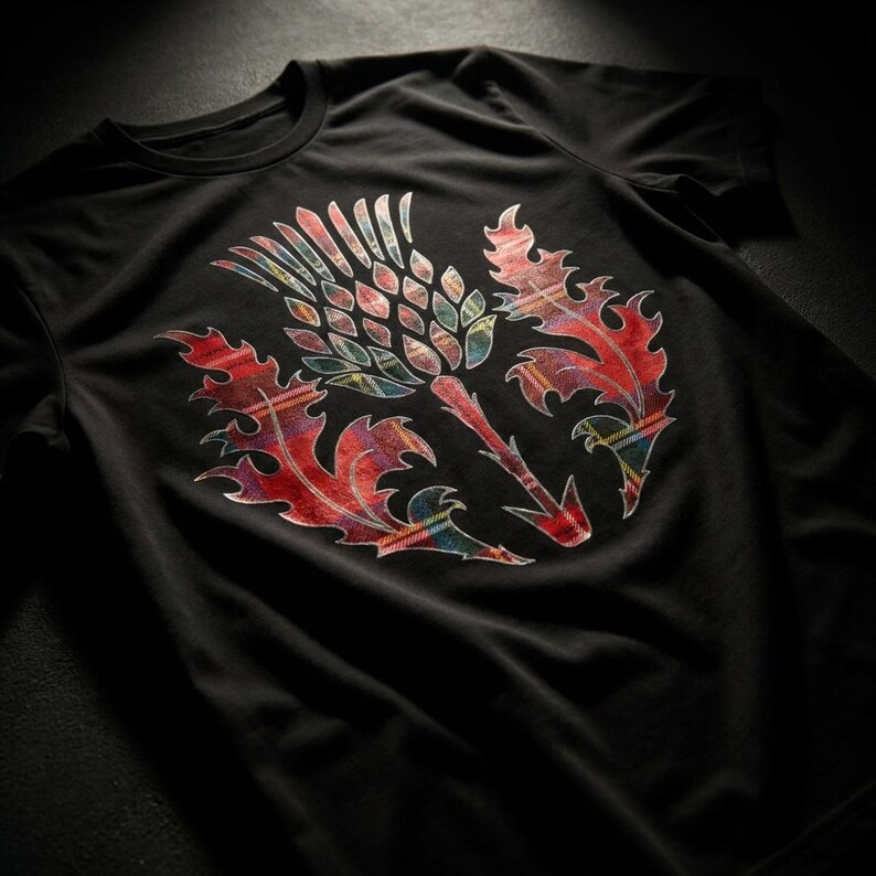 May include: A black short-sleeved t-shirt featuring a large graphic of a thistle plant on the chest. The thistle design is filled with a vibrant red, green, and blue tartan plaid pattern, contrasting against the solid black fabric of the shirt.