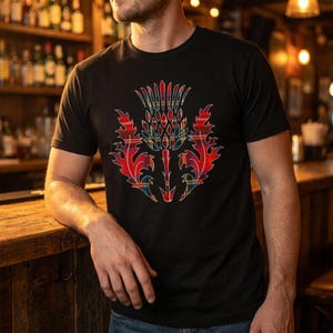 May include: A black short-sleeved t-shirt featuring a large, central graphic of a thistle plant. The thistle design is filled with a red, blue, and green plaid tartan pattern, creating a sharp contrast against the solid black fabric of the shirt.