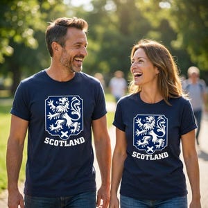 May include: A navy blue short-sleeved t-shirt featuring a white graphic of a rampant lion inside a shield shape above the text SCOTLAND. The design is centered on the chest of the garment, which has a crew neckline and a standard fit.