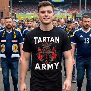 May include: A black t-shirt with the words "TARTAN ARMY" in white, and a red and green thistle design. The shirt is worn by a person walking in a crowd of people, many wearing blue and white scarves and shirts.