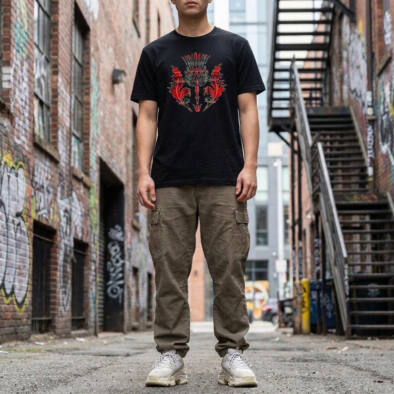 May include: A black short-sleeve t-shirt featuring a central graphic of a stylized red and green thistle design, paired with olive green cargo pants and white athletic sneakers. The t-shirt has a crew neckline and a standard fit.