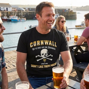 Puede incluir: Una camiseta negra de manga corta con el texto CORNWALL, WE'VE ONLY GOT ONE NEIGHBOUR, y WE DON'T LIKE THEM MUCH, con un gráfico de una calavera humana y huesos cruzados sobre un pequeño icono de escudo con un patrón dorado.