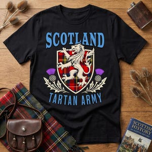 May include: A black short-sleeved t-shirt featuring a graphic print with the text SCOTLAND TARTAN ARMY in light blue. The design includes a central shield with a red, yellow, and blue tartan pattern, a cream-colored lion, and purple thistles.