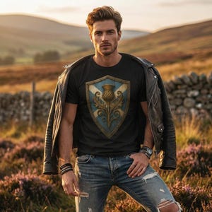 May include: A black t-shirt featuring a central graphic of a gold-rimmed shield with a blue and white saltire pattern and a detailed golden thistle emblem, worn under a black leather jacket with blue distressed denim jeans and wrist accessories.