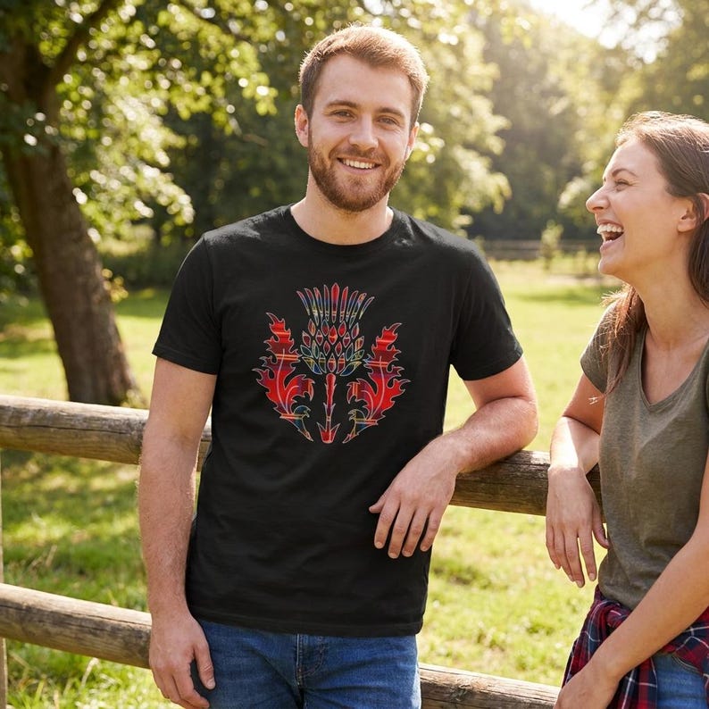 May include: A black short-sleeved t-shirt featuring a central graphic of a Scottish thistle. The thistle design is filled with a red, blue, and yellow tartan plaid pattern, creating a bold contrast against the solid black fabric of the shirt.