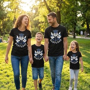 May include: A black short-sleeved t-shirt featuring a white and blue graphic of a Scottish thistle flower centered on the chest, with the word SCOTLAND printed in white capital letters in a semi-circular arc underneath the floral design.