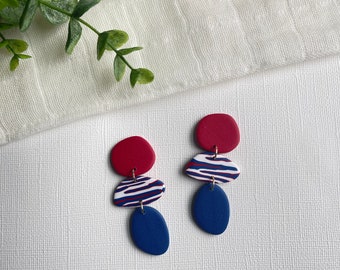 May include: A pair of handmade polymer clay earrings. Each earring features three stacked, organic shapes in red, blue, and white. The middle shape has a blue and white striped pattern. The earrings hang from small gold-colored hoops.