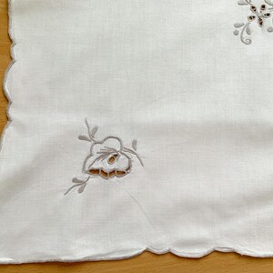 May include: A white rectangular textile with scalloped edges featuring delicate light gray floral embroidery and cutwork details. The fabric displays a smooth texture with symmetrical botanical patterns placed near the corners of the piece.