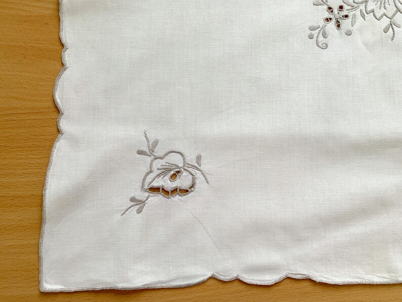 May include: A white rectangular textile with scalloped edges featuring delicate light gray floral embroidery and cutwork details. The fabric displays a smooth texture with symmetrical botanical patterns placed near the corners of the piece.