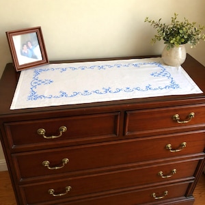 Può includere: Una tovaglia rettangolare bianca con ricami floreali blu. La tovaglia è esposta su una cassettiera in legno scuro, accanto a una foto incorniciata e un vaso con verde. La cassettiera ha maniglie in ottone.