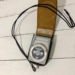 May include: A vintage light meter in a brown leather case with a black strap. The meter has a circular dial with numbers and text, including "Ленинград". The case is open, revealing the meter's face.