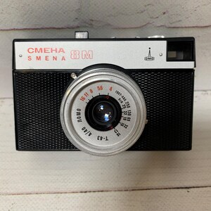 May include: A classic black and silver Smena 8M camera. The camera features a silver lens with black detailing and a black body. The text "СМЕНА SMENA 8M" is printed on the top. The lens has measurement markings.