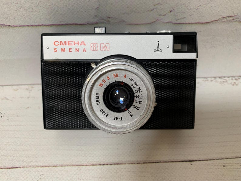 May include: A classic black and silver Smena 8M camera. The camera features a silver lens with black detailing and a black body. The text "СМЕНА SMENA 8M" is printed on the top. The lens has measurement markings.