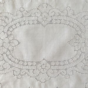 Può includere: Un tessuto bianco con un complesso ricamo a intaglio. Il design centrale è una cornice ovale composta da delicati motivi floreali e geometrici, creati con piccoli fori e sottili cuciture in filo bianco su tutta la superficie.