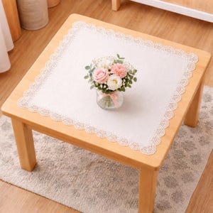 Può includere: Una tovaglietta quadrata in tessuto color bianco sporco con un delicato bordo in pizzo smerlato. Al centro della tovaglietta si trova un vaso in vetro trasparente contenente una composizione floreale di rose e garofani rosa e bianchi.