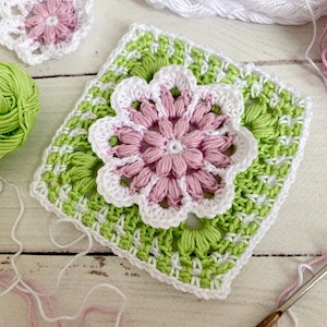 Puede incluir: Primer plano de artículos de ganchillo hechos a mano. Un granny square cuadrado con una flor rosa y blanca en el centro, rodeado de ganchillo verde y blanco. También se ven una pequeña flor de ganchillo y hilo.