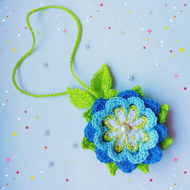 以下が含まれることがあります： 青、緑、白の花びらを持つ、かぎ針編みの花のネックレス。ネックレスは緑色の糸で作られており、花の中心に白い真珠のビーズが1つ付いています。