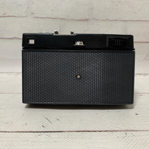 May include: A black vintage camera with a textured front panel and a silver-coloured top. The camera is rectangular with a small lens and various dials and buttons. The background is a white brick wall and a wooden surface.