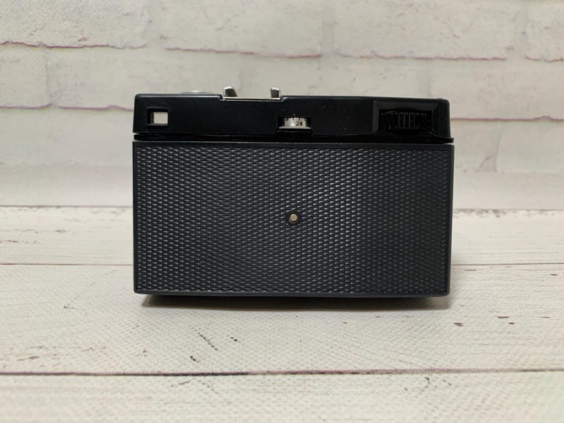 May include: A black vintage camera with a textured front panel and a silver-coloured top. The camera is rectangular with a small lens and various dials and buttons. The background is a white brick wall and a wooden surface.