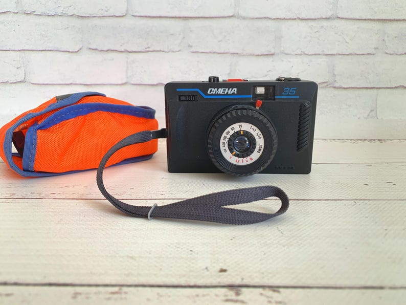May include: Black vintage camera with the text "Smena 35" and a dial. An orange and blue camera case and a grey strap are also present. The camera is on a light wood surface.