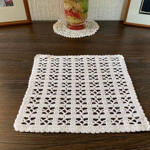 May include: A square, white, crocheted doily with a repeating floral pattern. The doily is placed on a dark brown wooden surface. A smaller, round doily is visible in the background, along with framed pictures and a decorative vase.