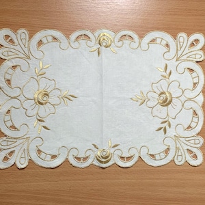 Puede incluir: Un tapete rectangular blanco con bordado floral dorado. El tapete presenta un borde festoneado y diseños florales intrincados, incluyendo flores grandes y pequeños acentos. El bordado dorado proporciona un contraste con la tela blanca.