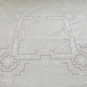 May include: A white square tablecloth featuring intricate white embroidery. The design includes a central geometric border with stepped corners, accented by small floral motifs and delicate leaf patterns stitched throughout the fabric.