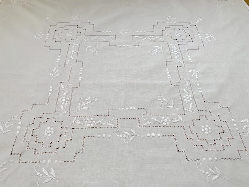 May include: A white square tablecloth featuring intricate white embroidery. The design includes a central geometric border with stepped corners, accented by small floral motifs and delicate leaf patterns stitched throughout the fabric.