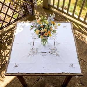 May include: A square white tablecloth featuring intricate brown floral embroidery and cutwork details. A clear glass vase sits in the center, filled with a colorful bouquet of yellow, orange, pink, and blue flowers, including roses and delphiniums.