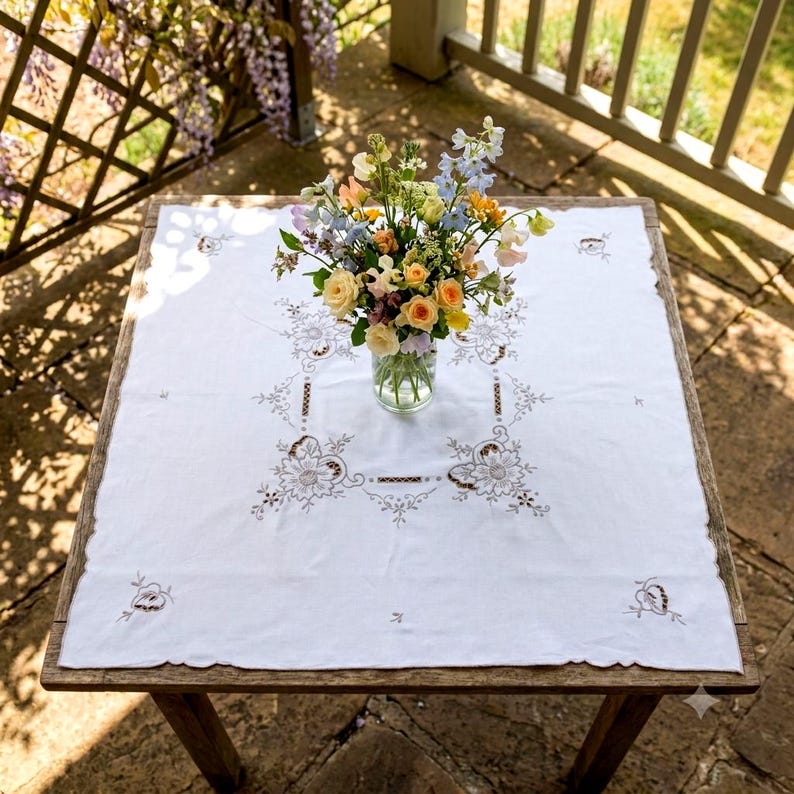May include: A square white tablecloth featuring intricate brown floral embroidery and cutwork details. A clear glass vase sits in the center, filled with a colorful bouquet of yellow, orange, pink, and blue flowers, including roses and delphiniums.