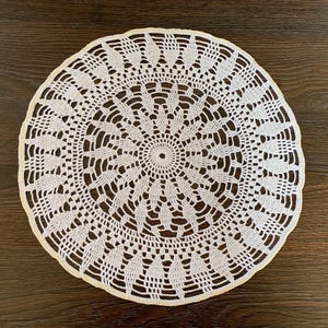 Op de afbeelding: Een witte, ronde doily met een ingewikkeld, opengewerkt haakontwerp. De doily heeft een centraal cirkelvormig patroon dat naar buiten uitstraalt, met een geschulpte rand. De doily is geplaatst op een donkerbruin houten oppervlak.