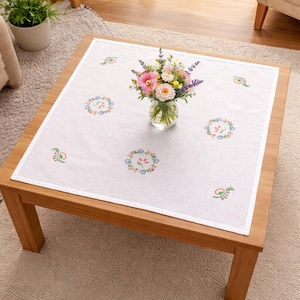 May include: A white square tablecloth with embroidered floral patterns and a white border. A glass vase holds a bouquet of pink, yellow, and purple flowers. The tablecloth is on a wooden coffee table.