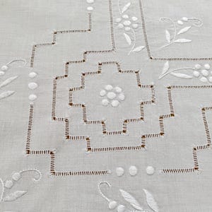May include: A white linen tablecloth featuring intricate white embroidery and cutwork. The design includes a central geometric stepped square pattern surrounded by delicate floral motifs, small circular accents, and linear stitching details.