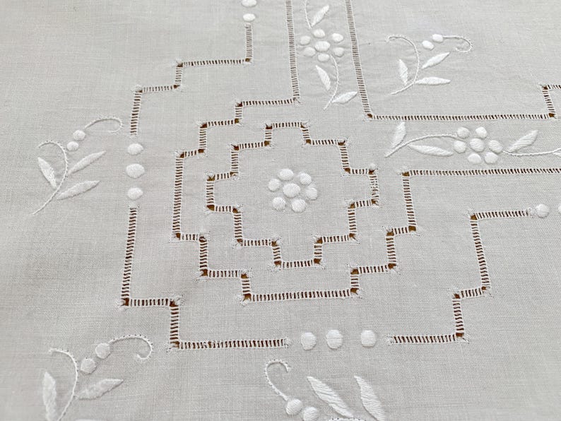 May include: A white linen tablecloth featuring intricate white embroidery and cutwork. The design includes a central geometric stepped square pattern surrounded by delicate floral motifs, small circular accents, and linear stitching details.
