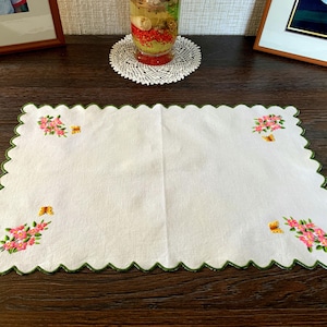 May include: A rectangular white placemat with a scalloped edge finished in dark green thread. Each of the four corners features embroidered pink flowers with green leaves and a small yellow butterfly accenting the floral clusters.