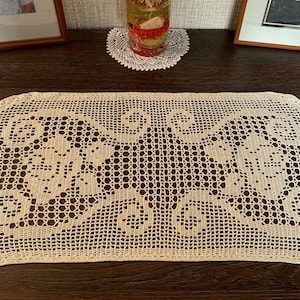 May include: Cream-colored, oval-shaped, crocheted table runner with a detailed openwork design. The runner features a repeating pattern of floral and scroll motifs, perfect for adding a vintage touch to any table setting.